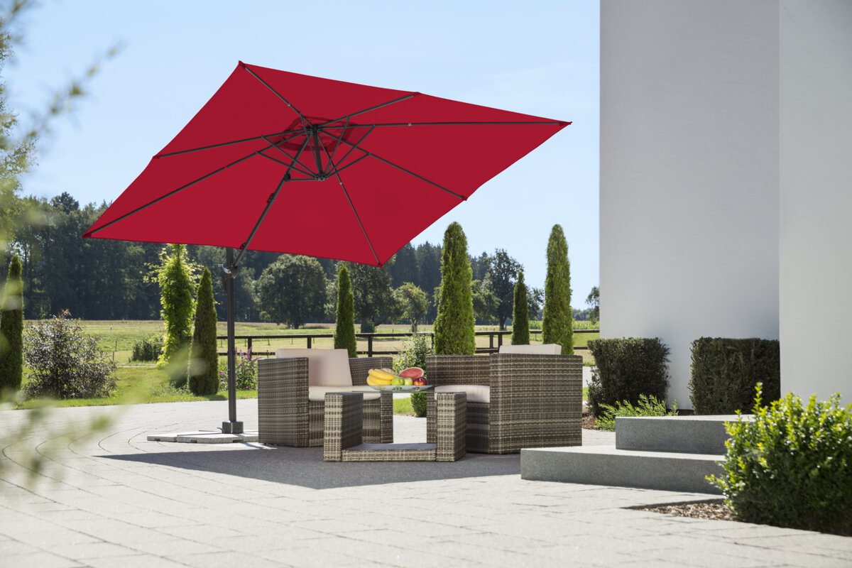 Schneider parasol Rhodos Junior in red, 270x270 cm, on a terrace with lounge furniture. A fruit bowl sits on the table. In the background are shrubs, a fence, a meadow with trees and a forest.