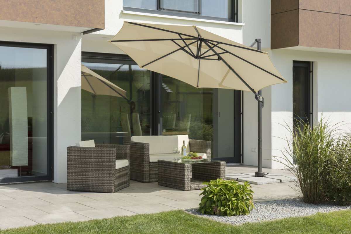 Schneider Rhodos Junior parasol in ecru, 300 cm in diameter, inclined, on a terrace with lounge furniture. A fruit bowl, a bottle and glasses are on the table. Small shrubs and a meadow are in the foreground.