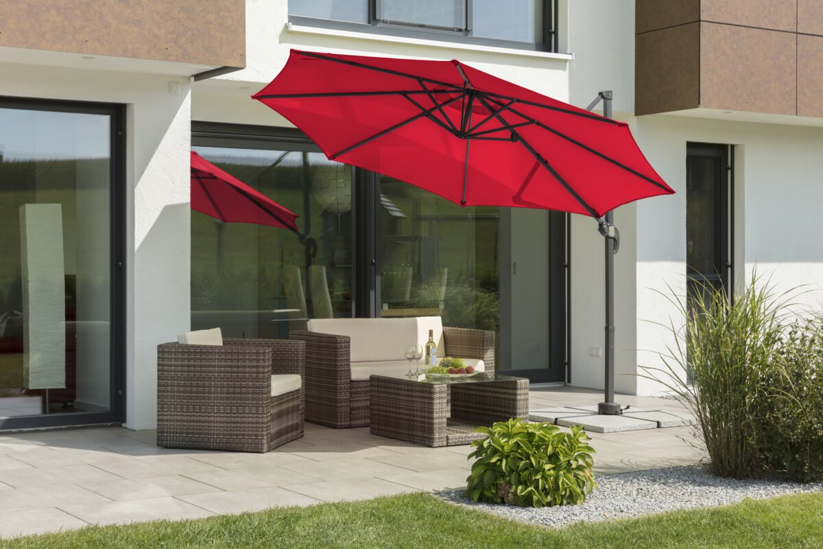 Schneider Rhodos Junior parasol in red, 300 cm in diameter, inclined, on a terrace with lounge furniture. A fruit bowl, a bottle and glasses are on the table. Small shrubs and a meadow are in the foreground.
