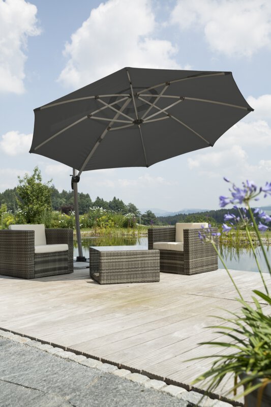 Schneider Rhodos Rondo parasol in anthracite, 350 cm in diameter, on a wooden terrace with lounge furniture by a natural swimming pool. In the foreground, a blooming purple flower and stone slabs, with a meadow, shrubs and forest in the background.