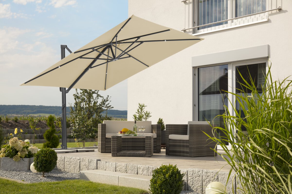 A Schneider Rhodos Smart parasol in ecru finish, 300x300 cm, on a terrace with lounge furniture below. A fruit bowl, a bottle and glasses are on the table. A stepped retaining wall made of concrete blocks is in the background. The terrace is surrounded by grass and shrubs.