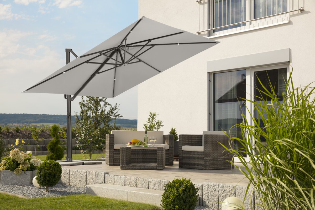 Schneider parasol Rhodos Smart in silver grey, 300x300 cm, on a terrace with lounge furniture below. A fruit bowl, a bottle and glasses are on the table. A stepped retaining wall made of concrete blocks is in the background. The terrace is surrounded by grass and shrubs.