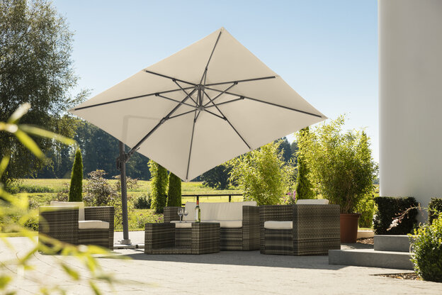 Schneider parasol Rhodos Twist in ecru, 300x300 cm, on a terrace with lounge furniture. A bottle and glasses are on the table. In the background are shrubs, a fence, a meadow with trees and a forest.