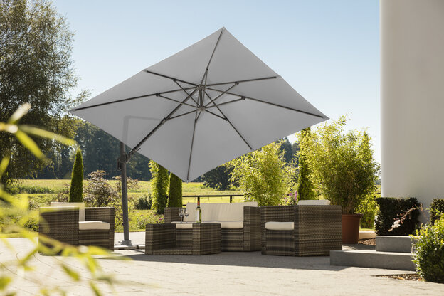 Schneider Sonnenschirm Rhodos Twist in Silbergrau, 300x300 cm, auf einer Terrasse mit Lounge-Möbeln. Auf dem Tisch befindet sich eine Flasche und Gläser. Im Hintergrund Sträucher, ein Zaun, eine Wiese mit Bäumen und Wald.