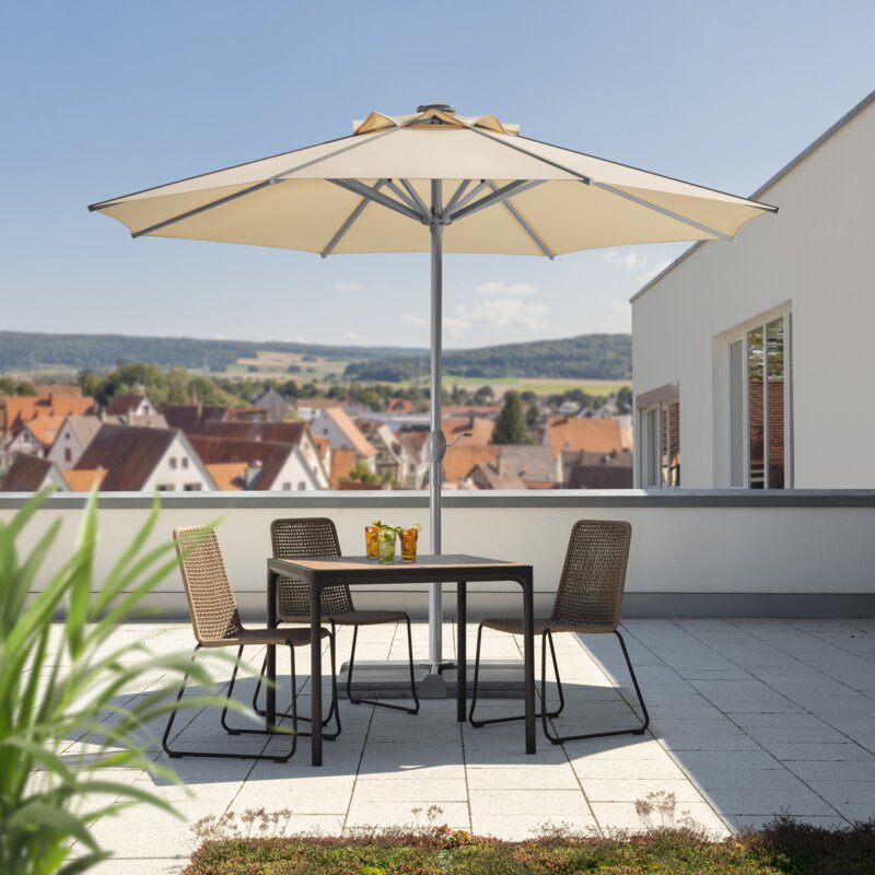 Schneider Rooftop parasol in ecru, 350 cm in diameter, on a roof terrace. Three chairs and a table are shaded by the parasol. Three cocktails are on the table. Numerous houses can be seen in the background. Behind the houses are meadows, hills and forest.