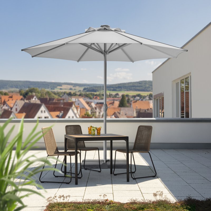 Schneider Rooftop parasol in silver grey, 350 cm in diameter, on a roof terrace. Three chairs and a table are shaded by the parasol. Three cocktails are on the table. Numerous houses can be seen in the background. Behind the houses are meadows, hills and forest.