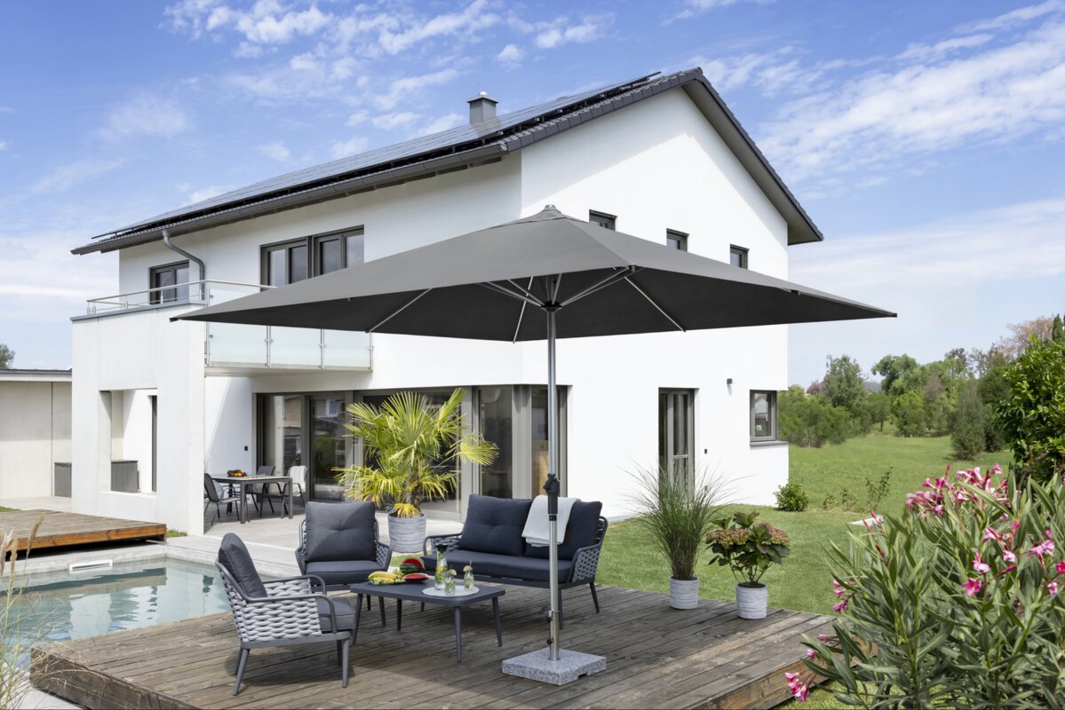 Schneider parasol Santorini in anthracite, 325x325 cm, on a wooden terrace in front of a residential building. Also visible on the terrace are two chairs and a bench around a small table with a carafe, two glasses and fruit. Surrounding the terrace are a pool and potted plants. In the background, a meadow and trees under a blue sky with light clouds.