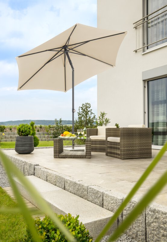 Schneider parasol Sevilla in ecru, 270 cm in diameter, on a terrace with lounge furniture below. A fruit bowl, a bottle and glasses are on the table. A stepped retaining wall made of concrete blocks is in the background. The terrace is surrounded by grass and shrubs.