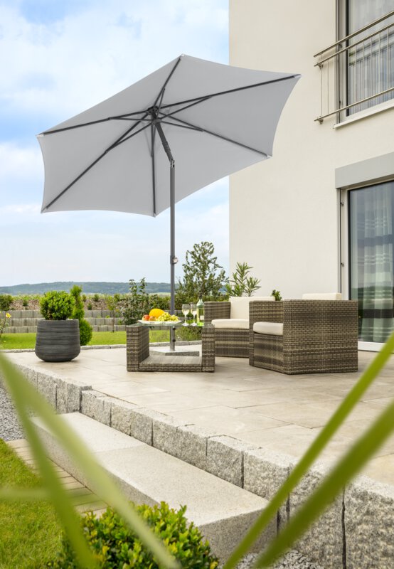 Schneider parasol Sevilla in silver grey, 270 cm in diameter, tilted, on a terrace with lounge furniture below. On the table is a fruit bowl, a bottle and glasses. In the background, a stepped retaining wall made of concrete blocks. The terrace is surrounded by grass and shrubs.