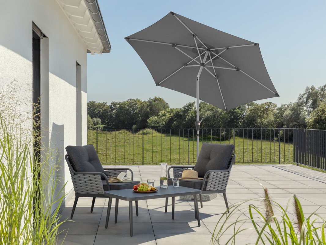 Schneider Sonnenschirm Solesta in Anthrazit, 270 cm Durchmesser, geknickt, auf einer Dachterrasse. Im Schatten des Schirms stehen zwei Stühle mit grauen Sitzpolstern und ein kleiner Tisch mit Obstschale, Karaffe und Gläsern. Auf den Stühlen ein aufgeschlagenes Buch und ein Hut. Im Hintergrund Wiese und Wald unter strahlend blauem Himmel.