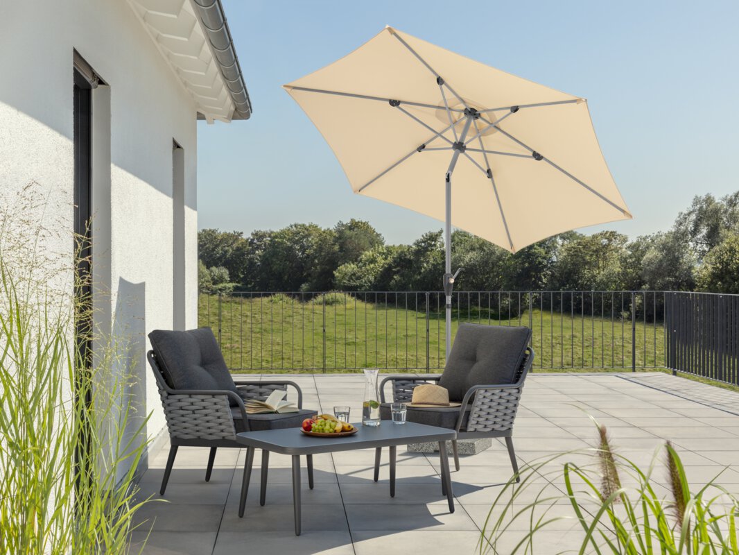 Schneider parasol Solesta in ecru, 270 cm in diameter, tilted, on a roof terrace. In the shade of the parasol are two chairs with gray seat cushions and a small table with a fruit bowl, carafe and glasses. On the chairs are an open book and a hat. In the background, a meadow and forest under a bright blue sky.