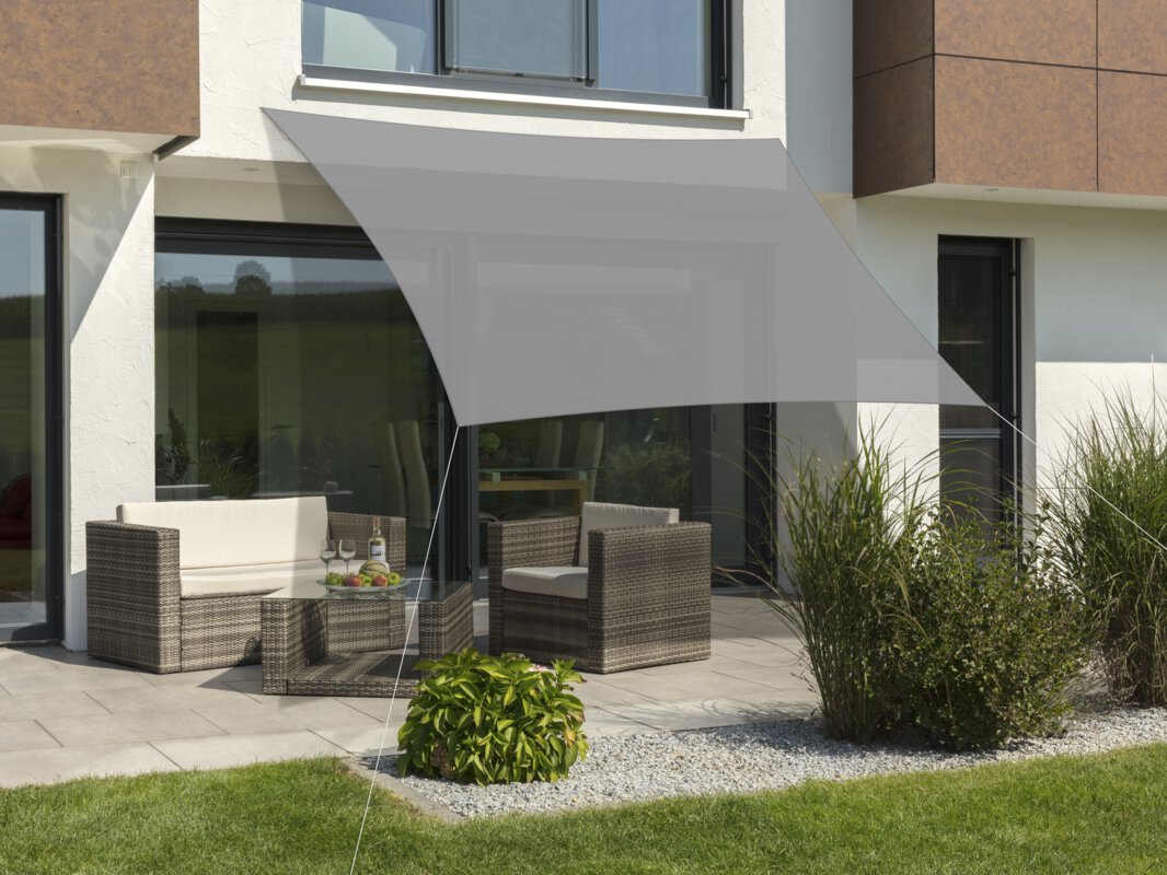 Schneider's Teneriffa sun sail in silver grey, 360x360 cm, stretched over a terrace with lounge furniture. A fruit bowl with a bottle and glasses sits on the table. Small shrubs and a meadow are in the foreground.