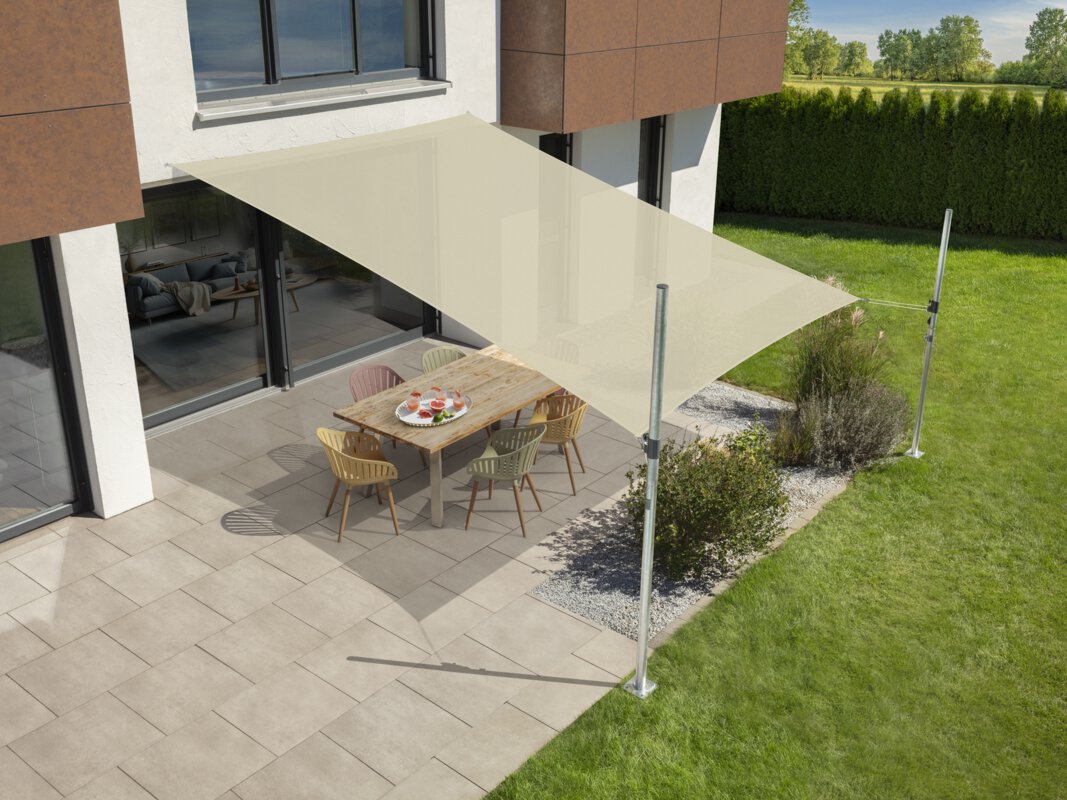 Schneider's Teneriffa sun sail in ecru, 500x360 cm, stretched over a terrace with garden furniture. A tray with drinks sits on the table. Small shrubs and a meadow are in the foreground. Behind a hedge, a meadow and trees.