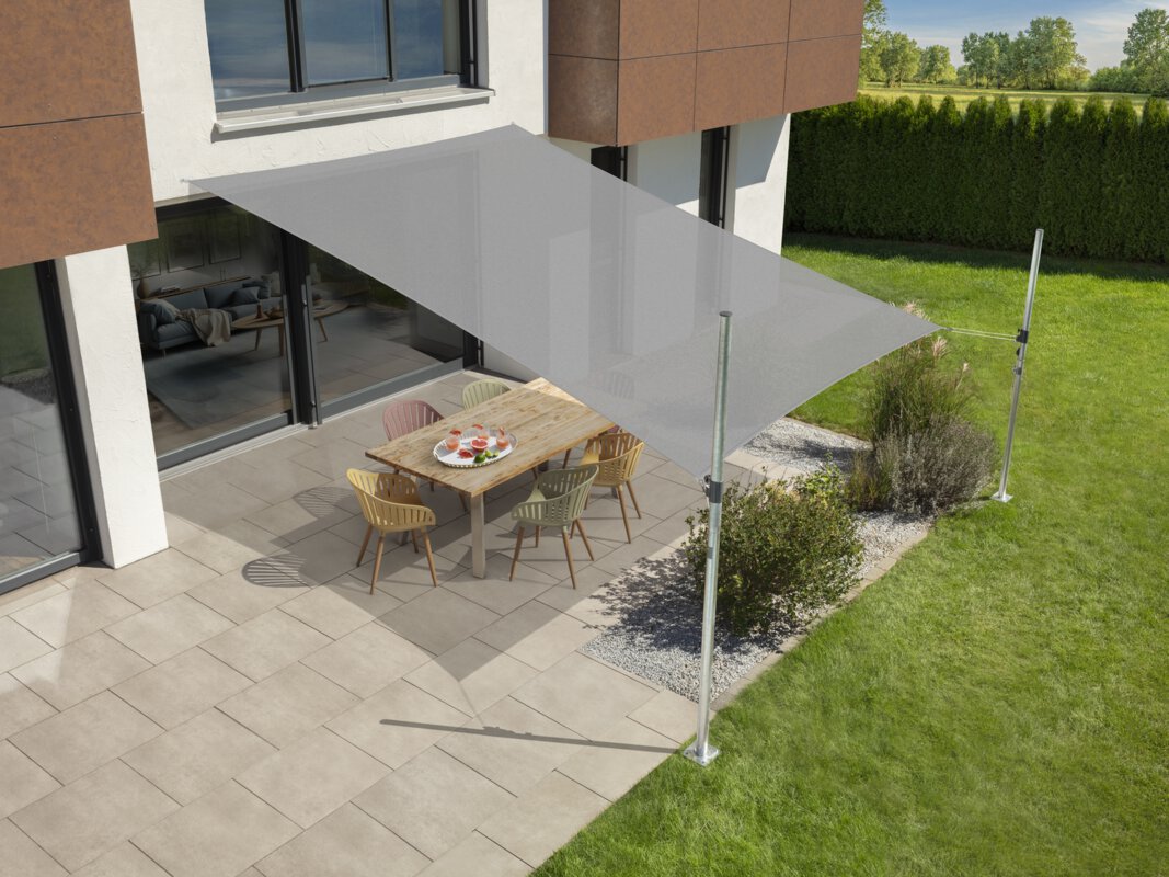 Schneider Sonnensegel Teneriffa in Silbergrau, 500x360 cm, gespannt über eine Terrasse mit Gartenmöbeln. Auf dem Tisch befindet sich ein Tablet mit Getränken. Im Vordergrund kleine Sträucher und Wiese. Hinter einer Hecke Wiese und Bäume.