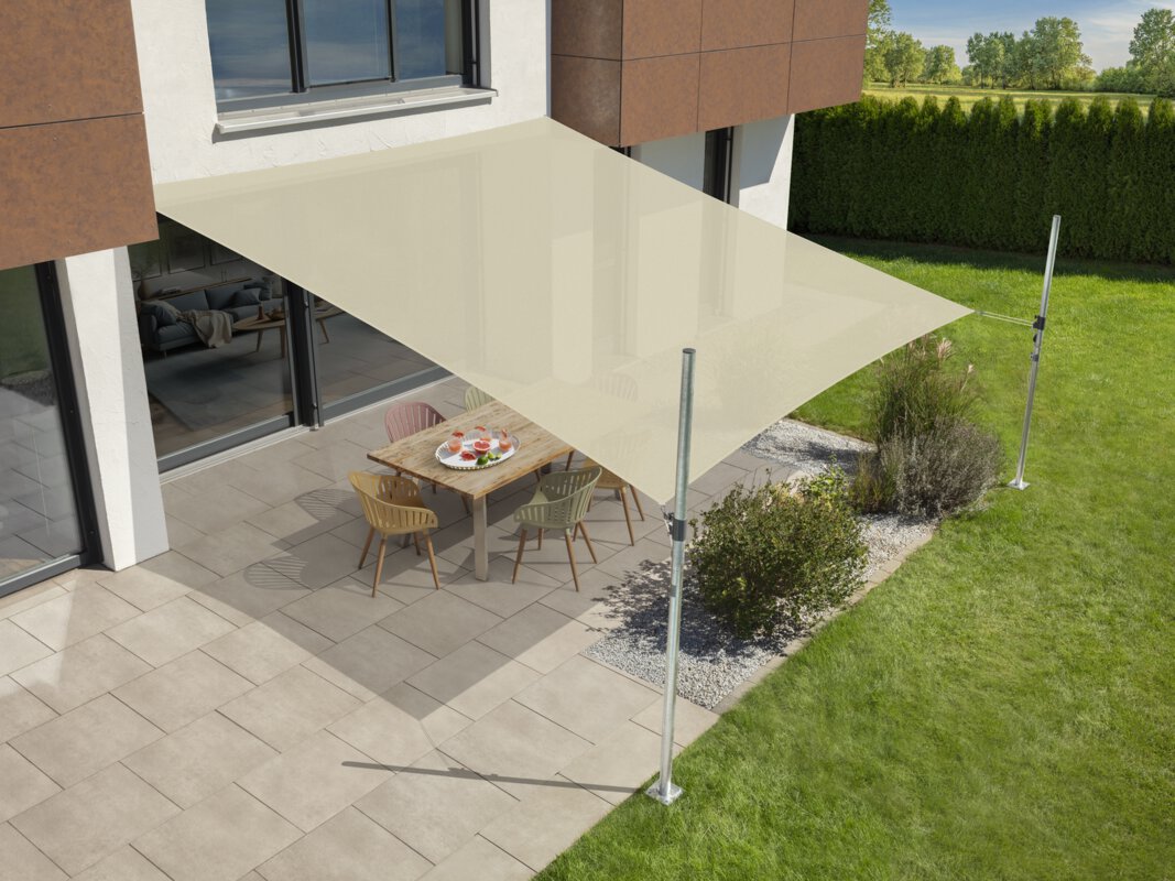 Schneider's Teneriffa sun sail in ecru, 500x500 cm, stretched over a terrace with garden furniture. A tray with drinks sits on the table. Small shrubs and a meadow are in the foreground. Behind a hedge, meadow and trees.