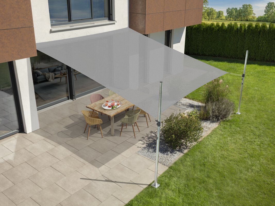 Schneider's Teneriffa sun sail in silver grey, 500x500 cm, stretched over a terrace with garden furniture. A tray with drinks sits on the table. Small shrubs and a meadow are in the foreground. Behind a hedge, a meadow and trees.