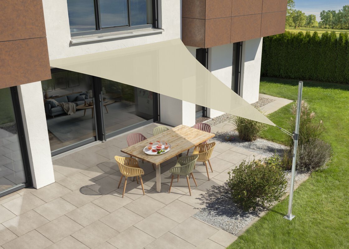 Schneider's Teneriffa sun sail in ecru, 500x500x500 cm, stretched over a terrace with garden furniture. A tray with drinks sits on the table. Small shrubs and a meadow are in the foreground. Behind a hedge, meadow and trees.