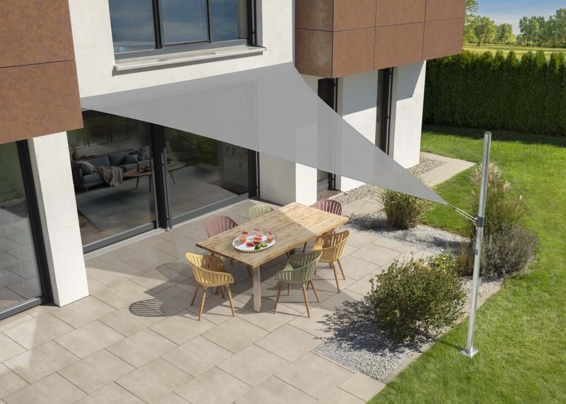 Schneider Sonnensegel Teneriffa in Silbergrau, 500x500x500 cm, gespannt über eine Terrasse mit Gartenmöbeln. Auf dem Tisch befindet sich ein Tablet mit Getränken. Im Vordergrund kleine Sträucher und Wiese. Hinter einer Hecke Wiese und Bäume.