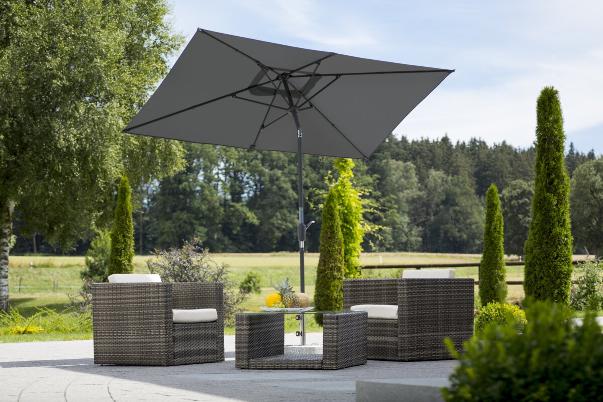 Schneider's Tunis parasol in anthracite, 270x150 cm, on a terrace with lounge furniture. A fruit bowl sits on the table. In the background are shrubs, a fence, a meadow with trees and a forest.