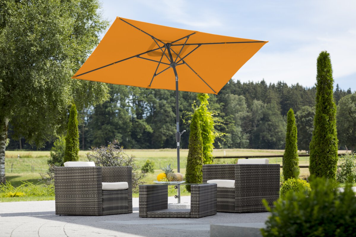 Schneider Tunis parasol in mandarin, 270x150 cm, on a terrace with lounge furniture. A fruit bowl sits on the table. In the background are shrubs, a fence, a meadow with trees and a forest.