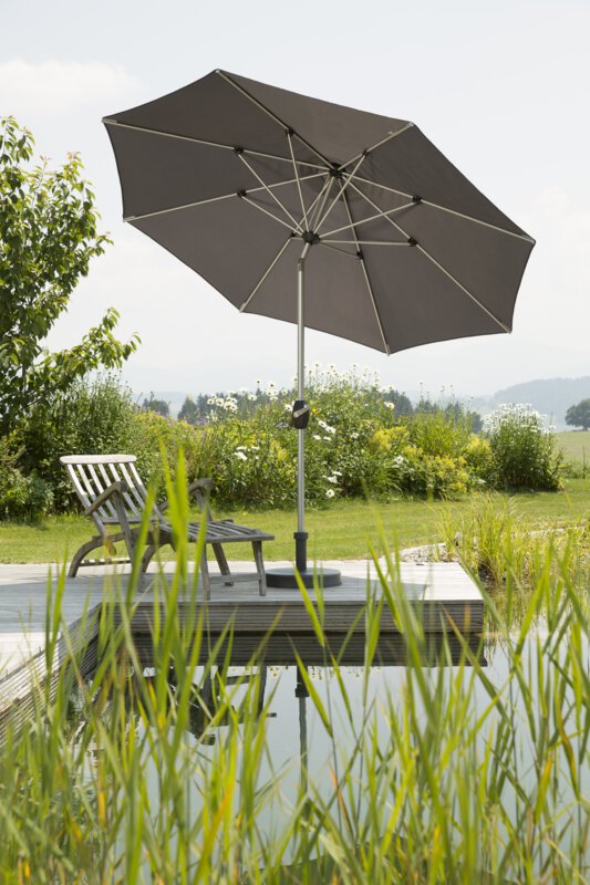 Schneider Venedig parasol in anthracite, 270 cm in diameter, on a wooden terrace by a natural swimming pool. A deck chair sits beneath it. Reeds are in the foreground and meadows and shrubs are in the background.