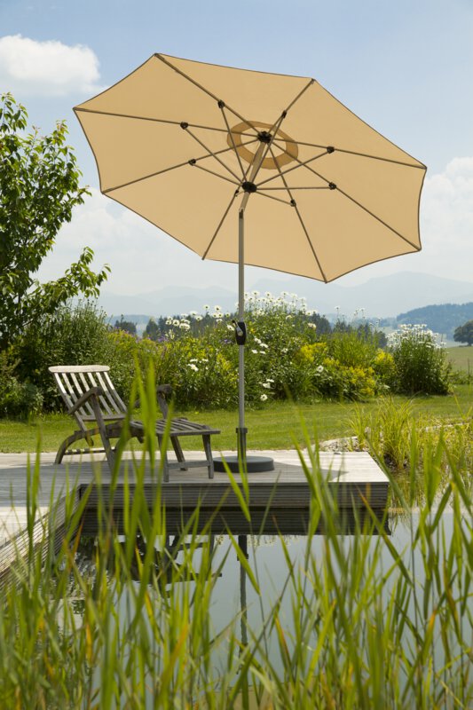 A Schneider Venedig parasol in ecru, 270 cm in diameter, placed on a wooden deck by a natural swimming pool. A deck chair sits beneath it. Reeds are in the foreground and meadows and shrubs are in the background.