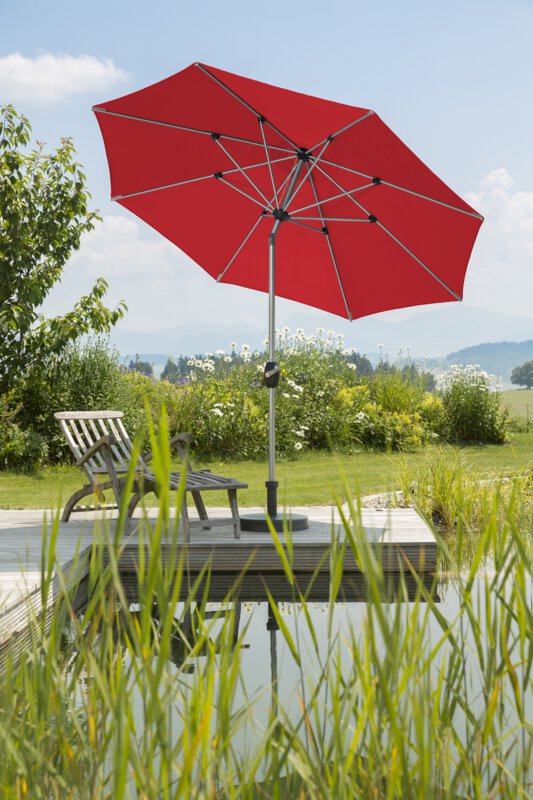 Schneider Sonnenschirm Venedig in Rot, 270 cm Durchmesser, auf einer Holzterrasse an einem Naturpool. Darunter steht ein Liegestuhl. Im Vordergrund Schilf, im Hintergrund Wiese und Sträucher.