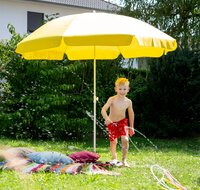 Schneider Sonnenschirm Ibiza mit 200 cm Ø in Goldgelb spendet angenehmen Schatten in einem Garten. Unter dem Schirm steht ein Kind neben einer Decke mit Kissen, im Vordergrund spritzt eine kleine Wasserfontäne – eine sommerliche, farbenfrohe Atmosphäre.