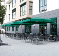 Drei Schneider Sonnenschirme Quadro 300×300 cm in Grün stehen zwischen Tischen und Stühlen in einem Gastronomiebereich und bieten entspannte, sonnengeschützte Sommerplätze, auch bei leichtem Regen.