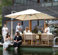 Grillparty auf der Terrasse mit Naturpool im Vordergrund, in dem zwei Personen ihre Füße eintauchen. Im Hintergrund dominiert der Sonnenschirm Rhodos Twist Woody in Natur, unter dem Personen mit Getränken sitzen und stehen.