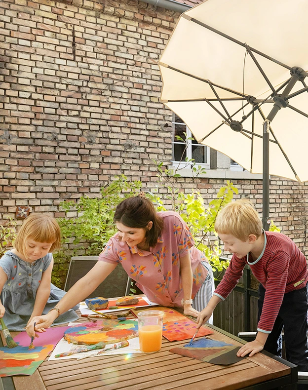 Schneider Schirme Salerno 350x150 cm in Natur im Einsatz -  öffnet und schließt selbst auf engem Raum. Eine Frau malt gemeinsam mit zwei Kindern an einem Tisch auf dem Balkon mit Wasserfarben unter dem Sonnenschirm Salerno 350x150 cm in Natur. Im Hintergrund ist die Hauswand eines gemauerten Hauses zu sehen.