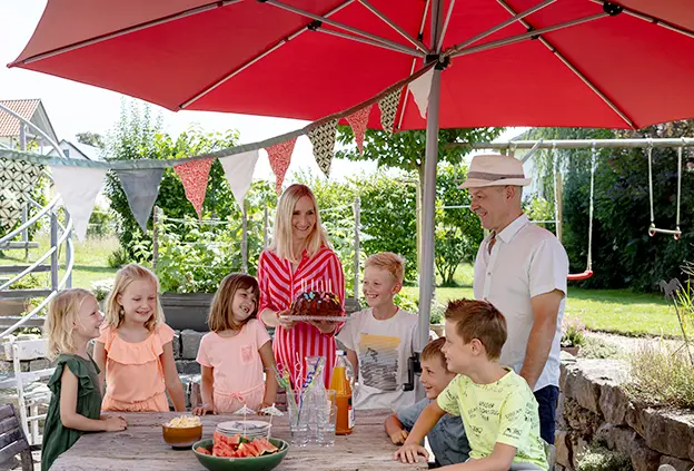 Sechs Kinder und zwei Erwachsene stehen unter dem Schneider Sonnenschirm Gemini an einem Tisch. Auf dem Tisch liegen Wassermelonenstücke, Chips und Teller. Außerdem stehen dort mehrere leere Gläser, eine Flasche Saft und eine Karaffe mit Wasser. Eine Frau hält einen Kuchen mit Kerzen in der Hand.