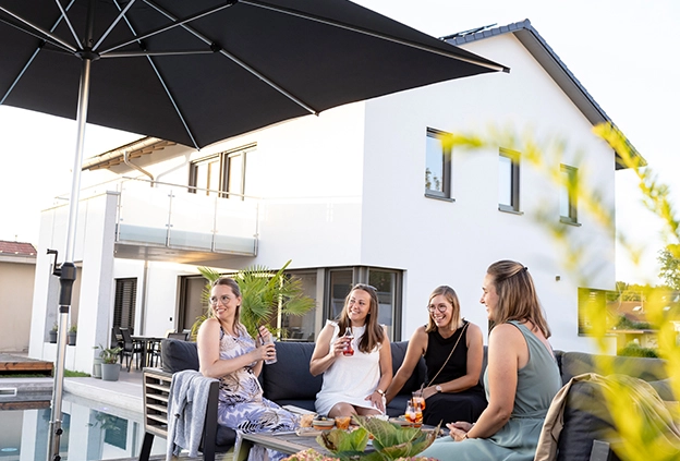 Vier Freundinnen sitzen auf einer Lounge am Pool und genießen gemeinsam Getränke und Snacks unter dem Schneider Sonnenschirm Santorini. Im Hintergrund ist ein modernes Wohnhaus zu sehen.