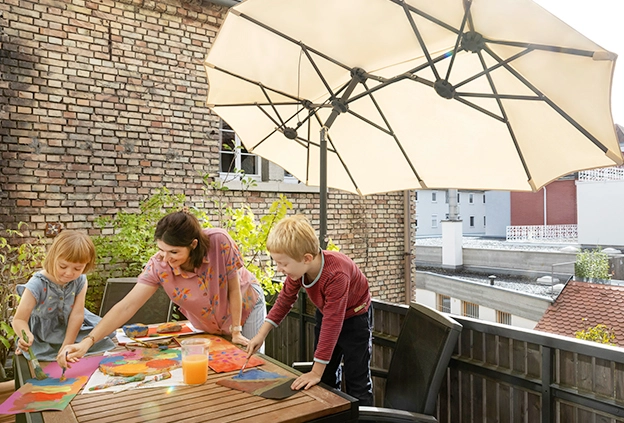 Eine Frau malt gemeinsam mit zwei Kindern an einem Tisch auf dem Balkon mit Wasserfarben unter dem Sonnenschirm Salerno 350x150 cm in Natur. Im Hintergrund ist die Hauswand eines gemauerten Hauses zu sehen.