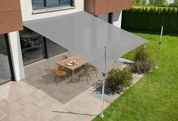 Silver grey Teneriffa sun sail by Schneider, stretched over a terrace and secured at multiple points. Under the sun sail are a table and chairs, surrounded by shrubs, lawn and hedges.