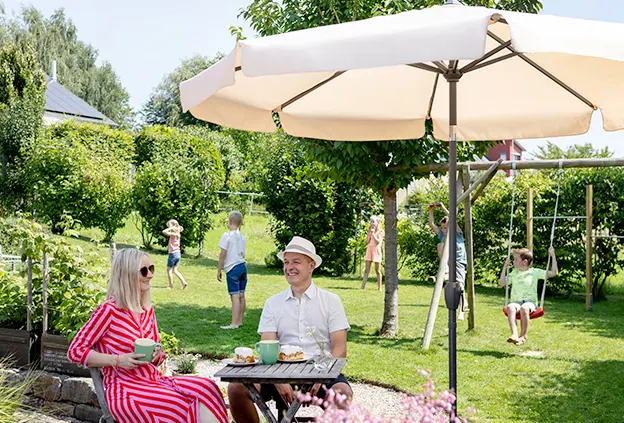 Großeltern sitzen an einem kleinen Tisch unter dem Schneider Orlando Sonnenschirm (Ø 270 cm) und genießen Kaffee und Kuchen, während Kinder im Hintergrund unter anderem auf einer Schaukel spielen.